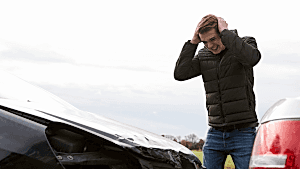 A distressed man clutches his head with his hands as he inspects the damage to the front of his car.